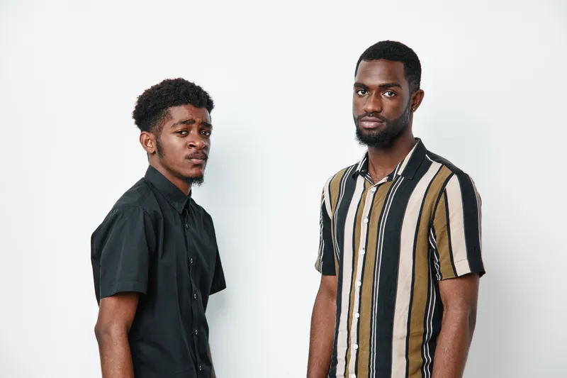 Two african american men with dark skin standing against white background wearing casual shirts looking at camera with serious expressions and confident posture.