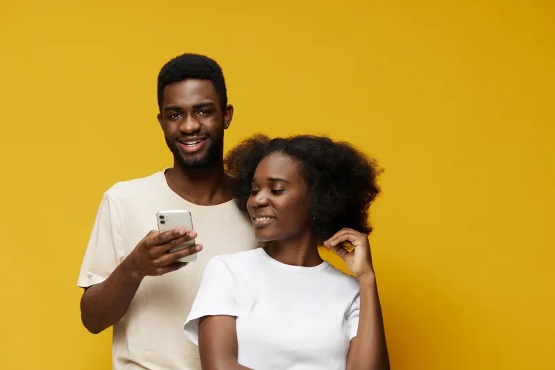 Two joyful young adults smiling together against a vibrant yellow background, showcasing modern casual attire and a playful relationship