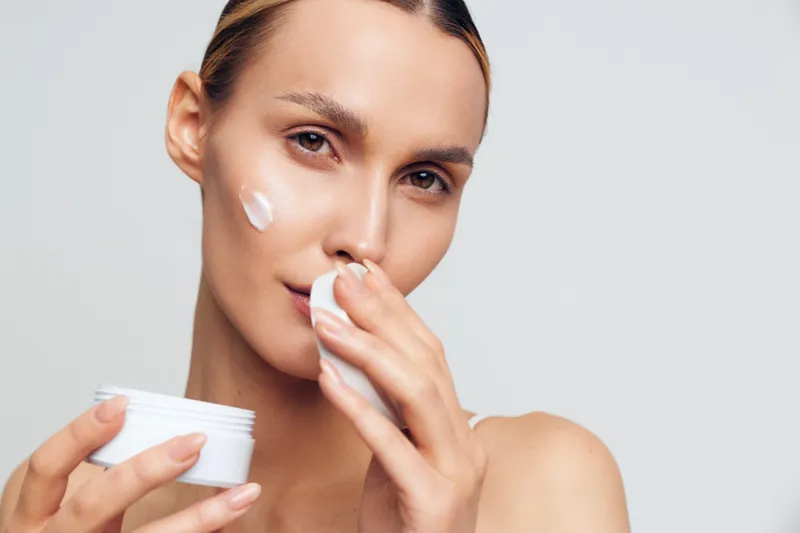 Young woman applying moisturizing cream on her face, clear skin, natural beauty, skincare routine, soft lighting, neutral background, close up portrait, healthy lifestyle