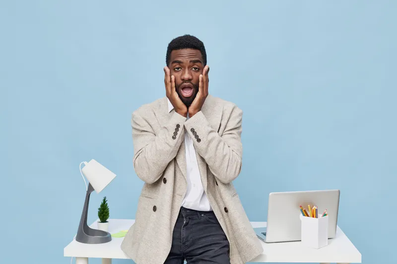 Frustrated young man with surprised expression, holding face with hands, standing at home office desk with laptop, lamp, and stationery on light blue background. People lifestyle concept