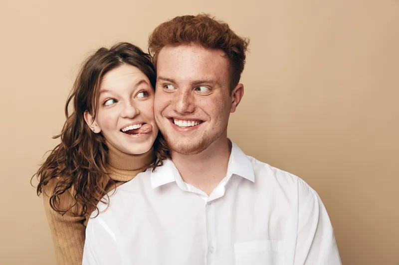 Happy young couple with curly hair smiling and looking at each other. Woman playfully sticks out tongue behind man in white shirt. Concept of love, friendship, and joyful relationship.