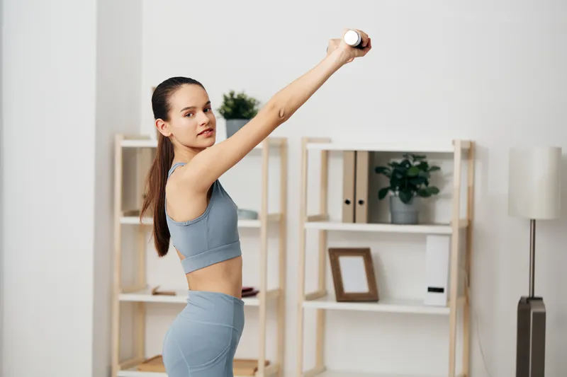 Woman fitness athlete performing an overhead dumbbell press in a clean home setting, showcasing a focused workout, strength training, balance, and form during a practical home exercise session.