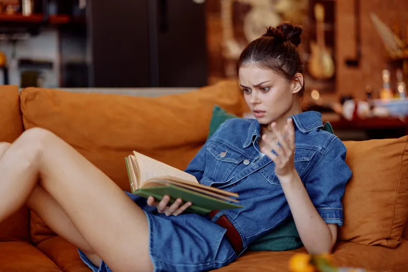 Женщина в джинсовом платье сидит на диване, скрестив ноги, и читает книгу в уютной домашней обстановке
