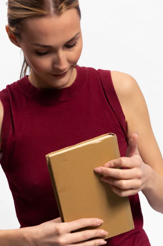 Young woman in red sleeveless top holds a closed book and looks down at it thoughtfully. Studio shot with white background. Concept of reading, education, and concentration.