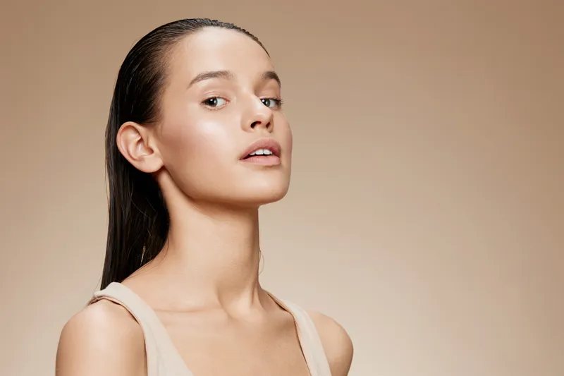 Natural beauty portrait of a young woman with long hair against a soft beige background, showcasing a healthy glow and minimal makeup for a fresh summer look