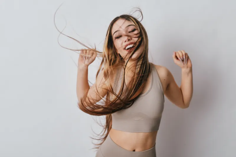 Young woman dancing with long hair flying, smiling and wearing beige sportswear on white background. Energetic movement, happiness and casual fitness style with joyful expression.