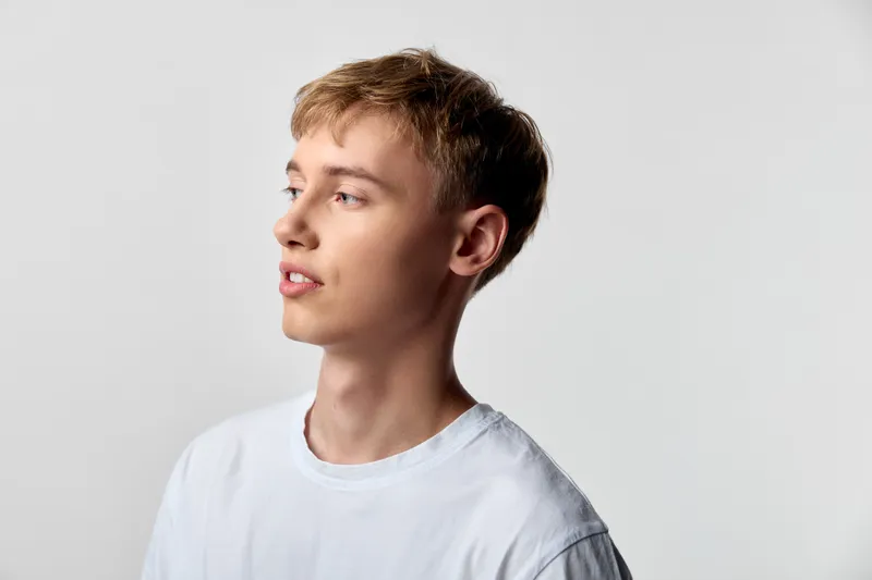 Young man in side profile looking away, wearing a white t-shirt in a clean studio with soft lighting and a neutral background, conveying casual style and contemporary fashion mood