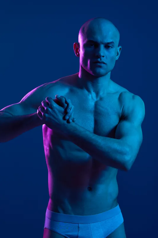 A toned man poses dramatically under vibrant blue and pink lighting, showcasing strength and confidence. The vivid colors enhance the mood and highlight his physique.