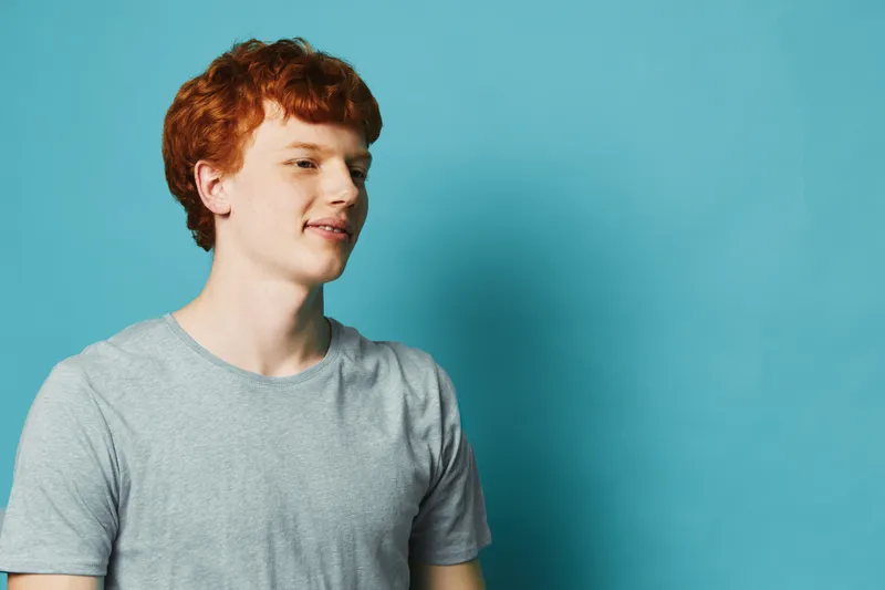 Young man with red hair wearing gray t shirt standing against blue background looking to side with calm expression and relaxed posture in studio setting