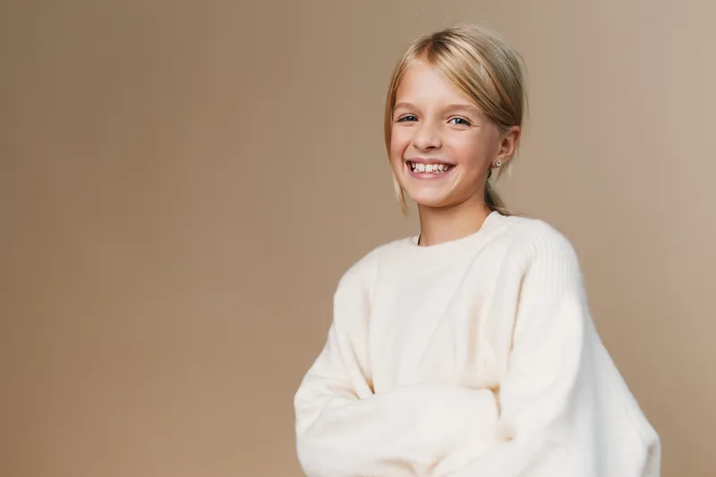 Smiling woman in a soft white sweater stands against a warm beige backdrop, radiating relaxed confidence and approachable charm ideal for lifestyle, fashion, and wellness imagery.