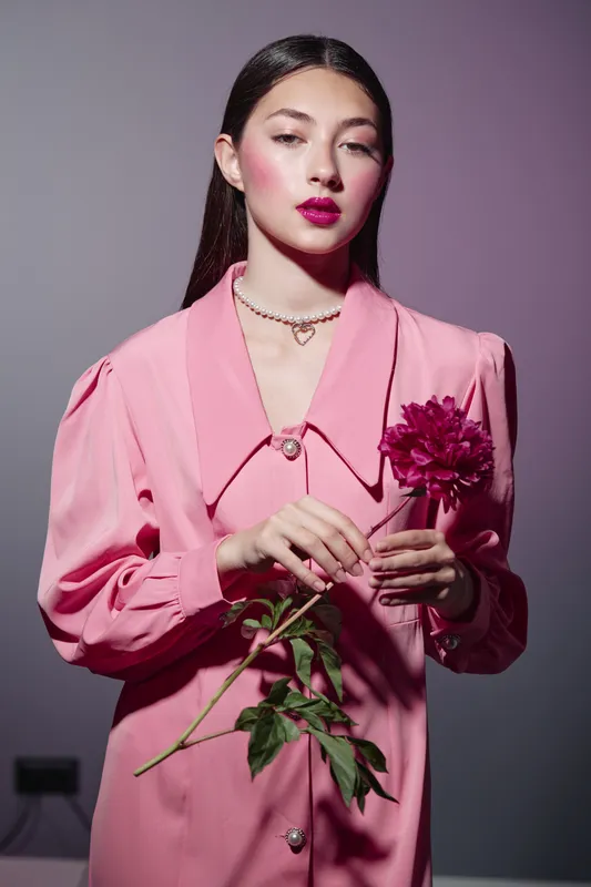 Young woman in pink vintage-style dress holds a magenta flower, posing creatively. She has bold makeup and Gatsby wave hairstyle, studio lighting highlights fashion and beauty concept.