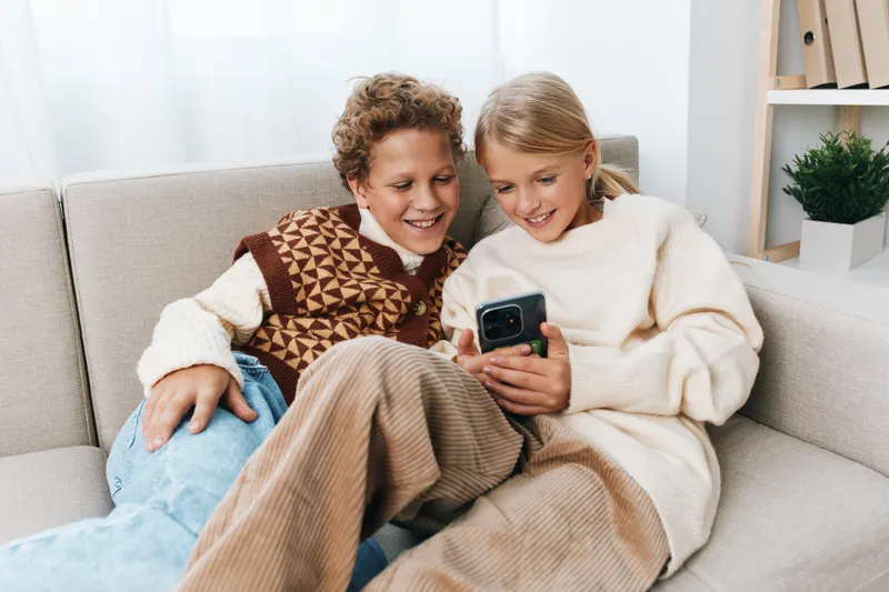 Two smiling children sit close on a sofa, sharing a moment as they look at a smartphone together, wearing cozy sweaters and relaxed at home in a bright living room.