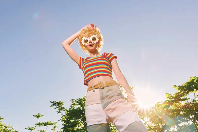 Young woman wearing a striped crop top and denim shorts stands under bright sun, a flowered hat and sunglasses completing a carefree summer vibe on a warm day.