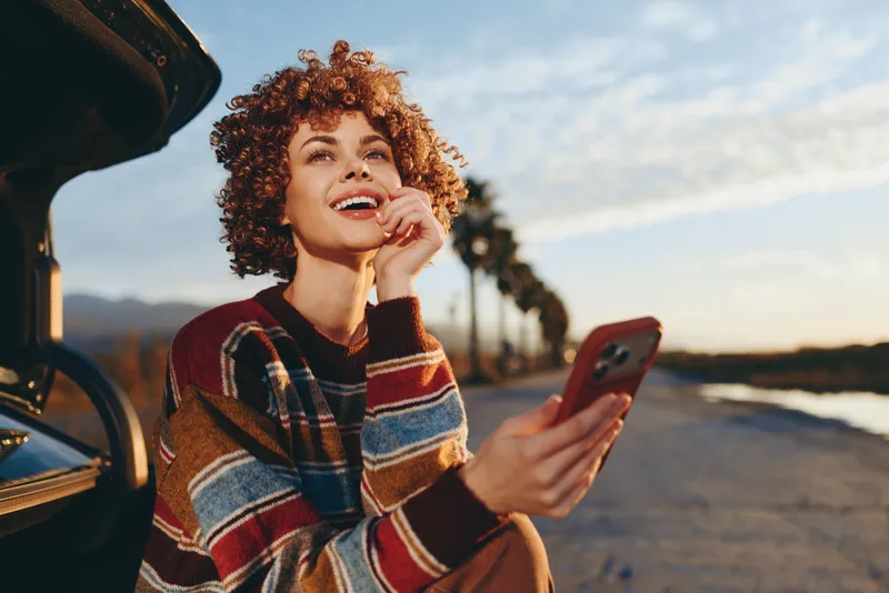 Woman lifestyle portrait outdoors by car on road, smiling while holding smartphone near her ear, striped sweater and curly hair, bright sky, blurred trees and street in distance