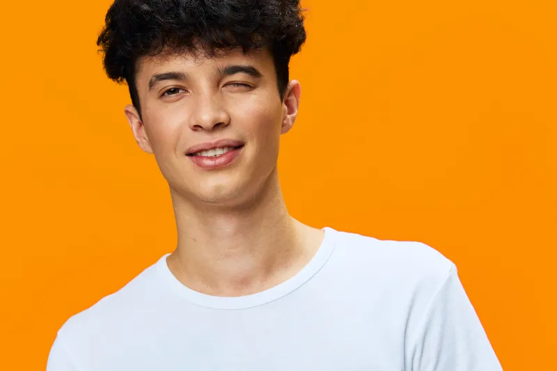 smiling man winking with curly hair, wearing white t-shirt, on vivid orange background, young adult, casual portrait expression
