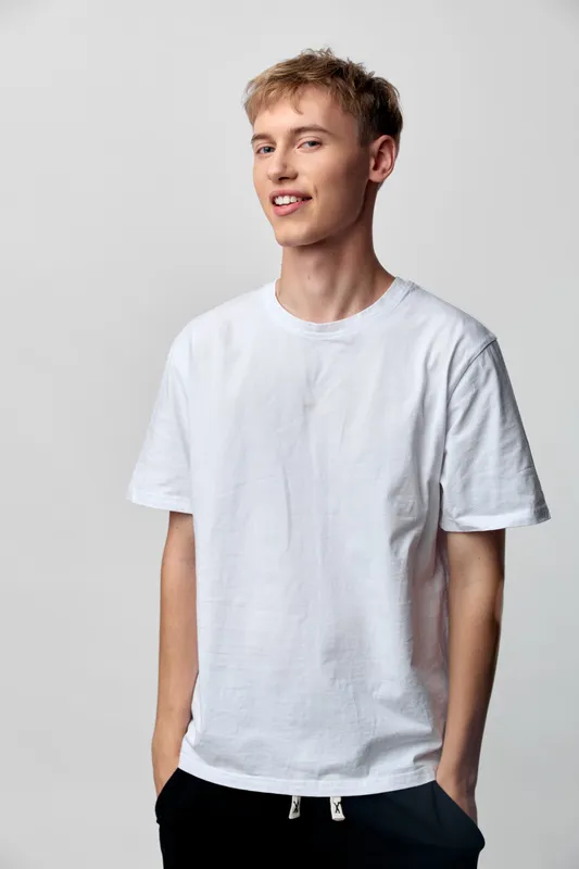 Man in white t shirt stands in a studio with relaxed posture, soft lighting, casual fashion, subtle smile, and simple clothing, creating an approachable everyday mood in clean minimalist setting