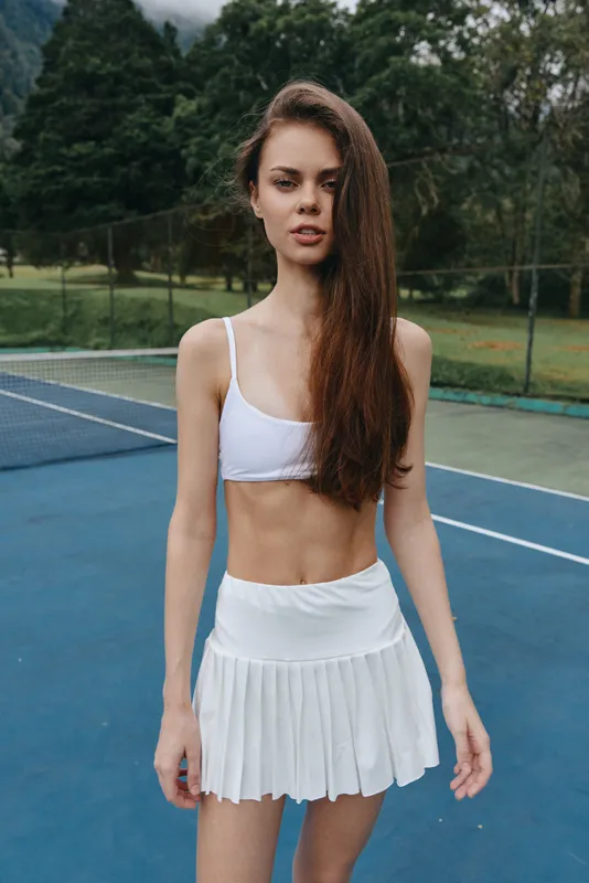 Young athletic woman in a white tennis outfit poses confidently on the court, showcasing a fit physique and determination