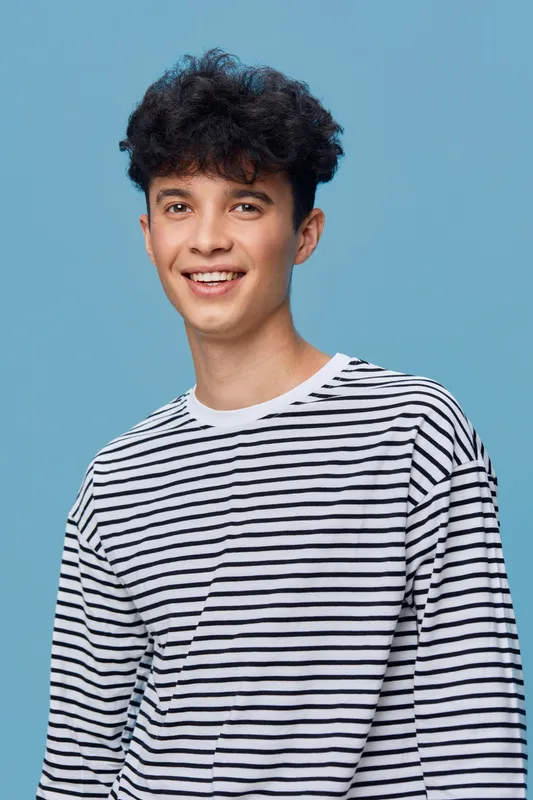 young man smiling in striped shirt with curly hair standing against light blue background, casual style and relaxed mood in bright studio setting