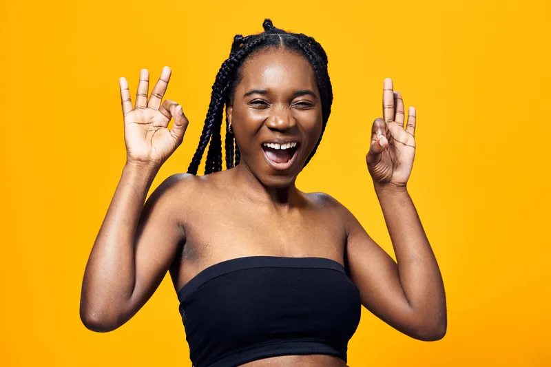 Joyful young woman with braided hair wearing a black crop top, smiling against a bright yellow background, conveying happiness and positivity