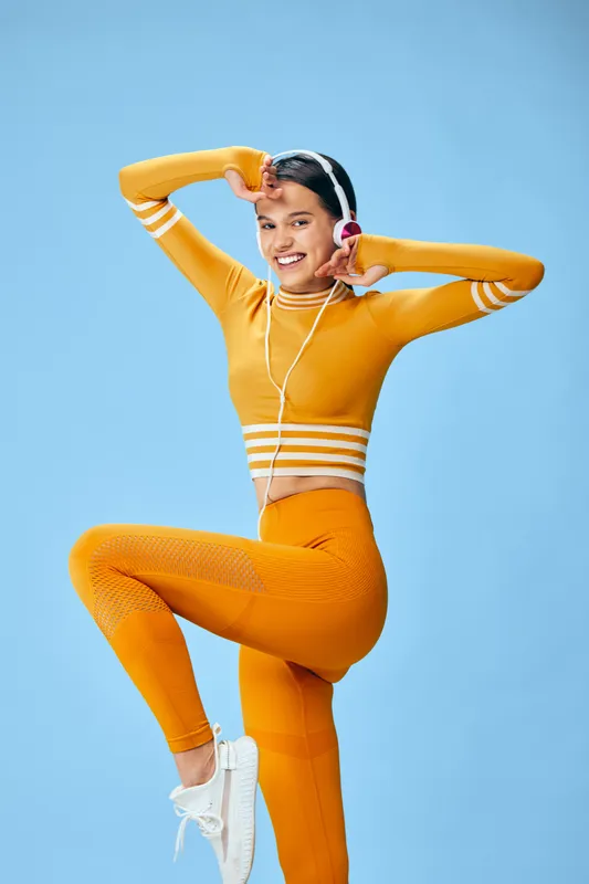 Woman in bright orange fitness gear performing a dynamic pose against a vivid blue studio backdrop, wearing headphones and athletic sneakers, conveying energy, movement and an active lifestyle.