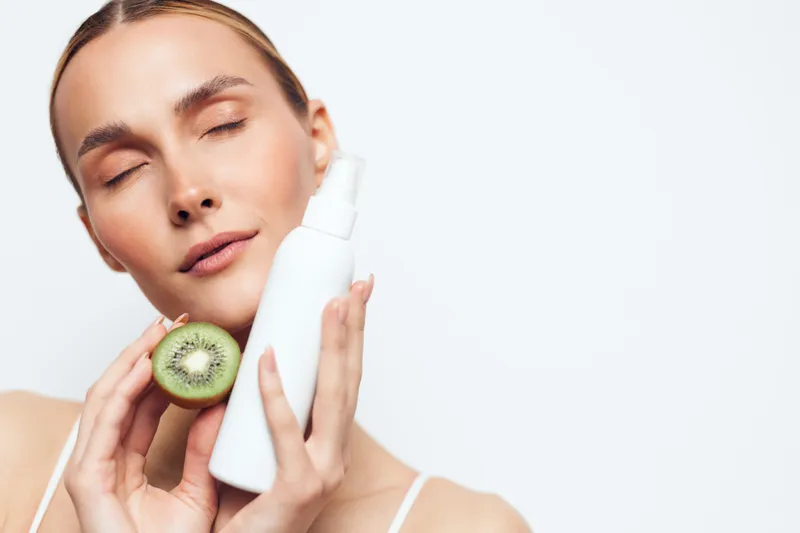 Young woman with clear skin holding a kiwi fruit and a white cosmetic bottle, eyes closed, minimal makeup, skincare concept, fresh natural beauty, clean background