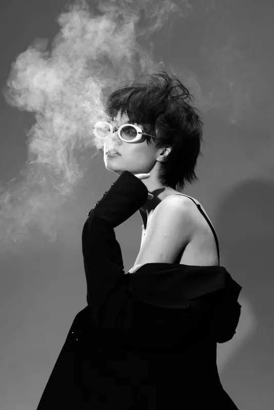 Black and white portrait of a stylish woman wearing sunglasses exhaling smoke with a moody atmosphere and retro fashion style.