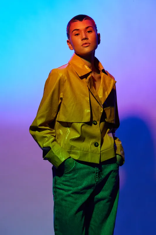 Young woman posing confidently in a shiny yellow jacket and green pants against a vibrant gradient background under colored lighting for a modern fashion look.