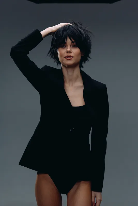 Stylish woman with short black hair wearing black blazer posing confidently against grey background.