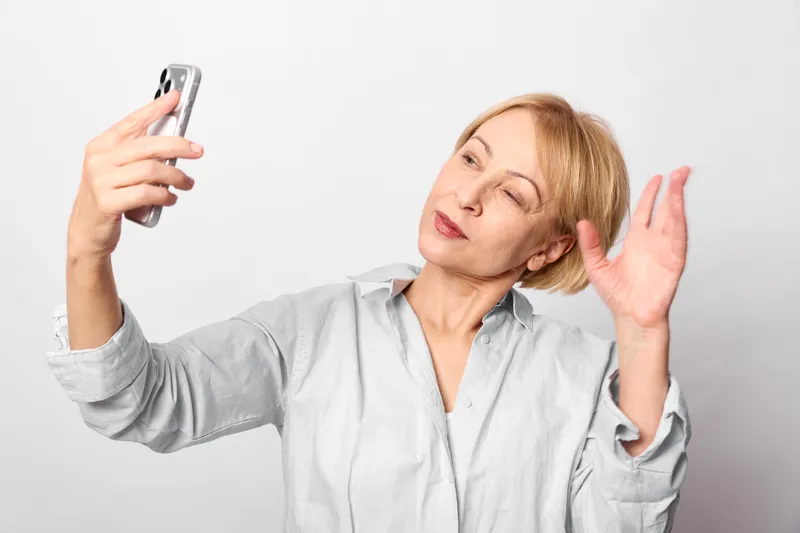 Woman holds a smartphone for a selfie, making a listening gesture beside her ear, indoor studio with clean white background, expressive portrait, natural light, relaxed pose soft