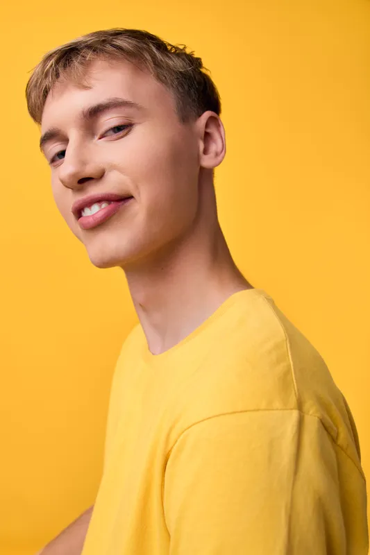 Man in a yellow shirt stands against a bright yellow backdrop, smiling with a relaxed pose. Studio closeup highlights smooth skin, casual vibe, and confident expression for fashion or lifestyle use.