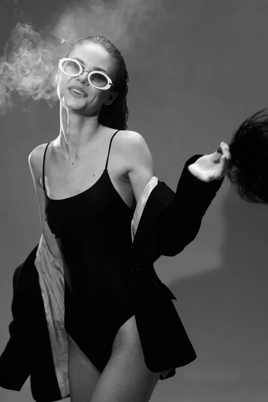 Smiling woman in sunglasses and black outfit exhaling smoke in black and white stylish portrait.
