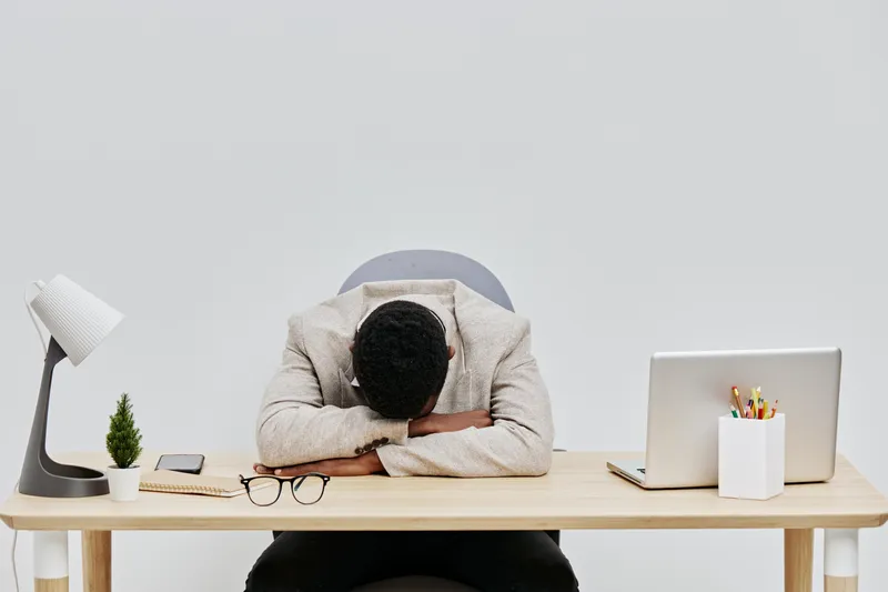 Overworked tired man in office resting head on desk with laptop and eyeglasses, indoor workspace with minimal decor, stress and fatigue concept, monochrome tones, empty background.