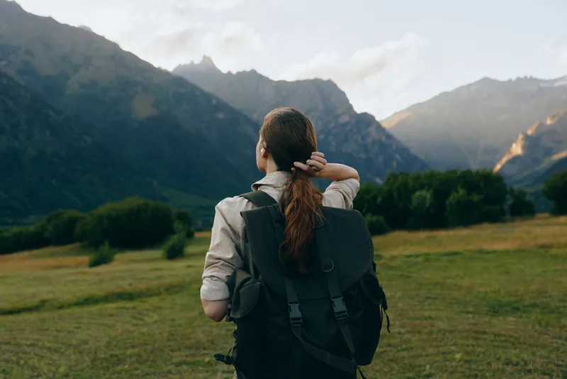 Young woman with a backpack gazes toward distant rugged mountains, ready for a hike, standing on a green meadow under a bright blue sky calm valley