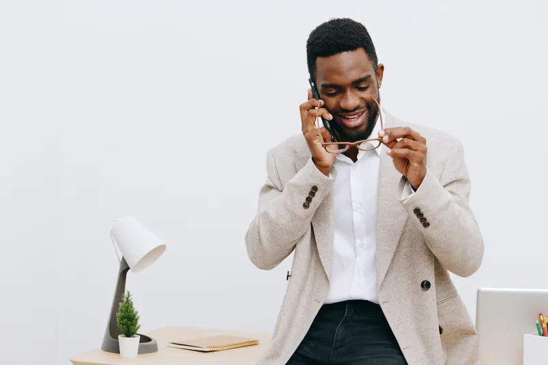 Modern professional man talking on mobile phone in office setting, smiling, wearing smart casual blazer and white shirt, standing near desk with lamp, plant, and laptop, light background, lifestyle co
