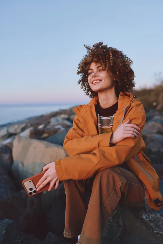 Smiling woman with curly hair in an orange jacket sits on rocks by the sea, holding a smartphone. Outdoor lifestyle at sunset, gentle ocean breeze, soft natural light for travel moments