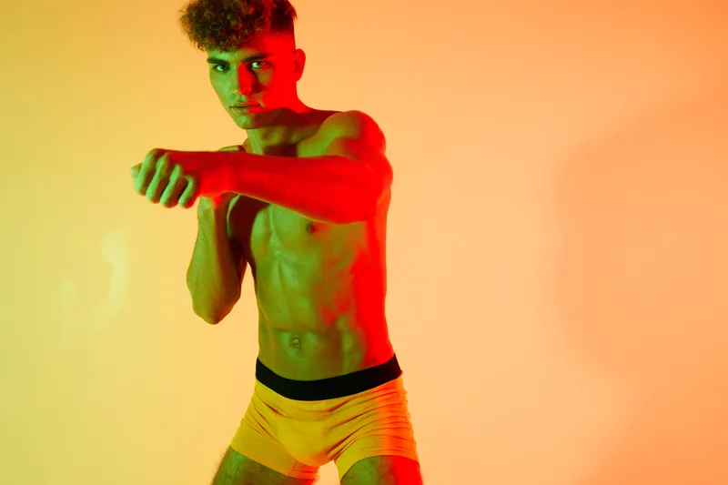 Muscular man posing shirtless in boxing assertive stance with yellow shorts and colorful lighting, training fighting passion workout at studio gym background strength exercise.