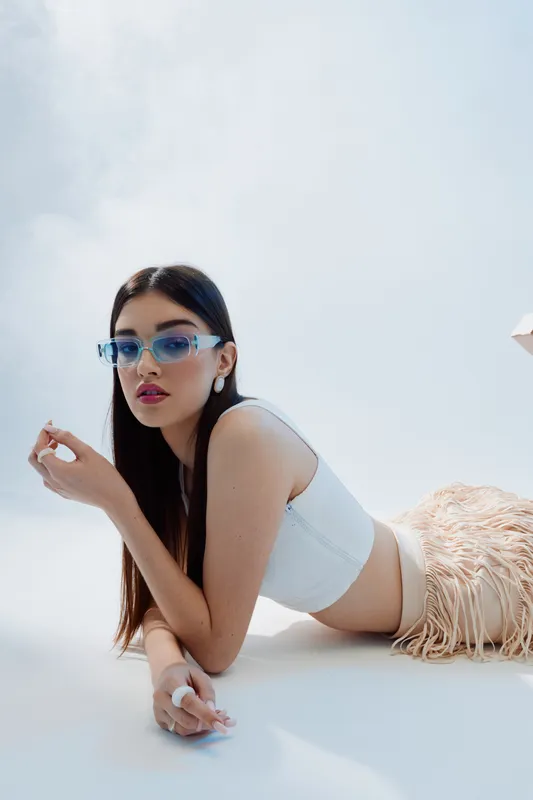 Young woman in white top and beige skirt poses lying down wearing blue sunglasses. Minimalist studio shot with soft lighting. Concept of woman beauty fashion and modern style.