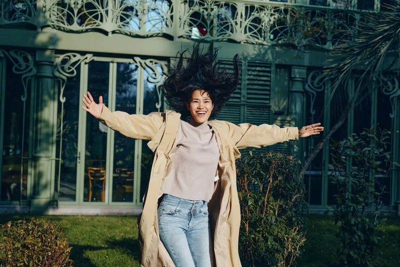 Joyful woman wearing a beige coat leaps outdoors beside an ornate gate, casual style and carefree energy, spring evening light, expressive movement in a residential garden setting