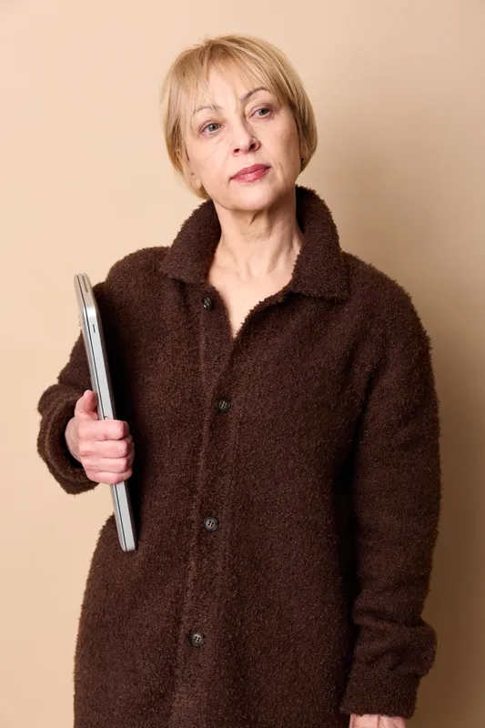 Senior woman in brown coat holding a tablet, studio portrait on beige background. Technology and communication concept with confident calm expression, soft studio lighting, warm mood.