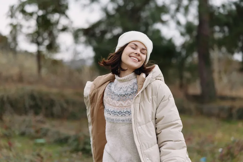 Woman in a warm beige jacket and knit sweater smiles outdoors in a park, wearing a beanie, capturing a casual winter portrait with soft natural light.