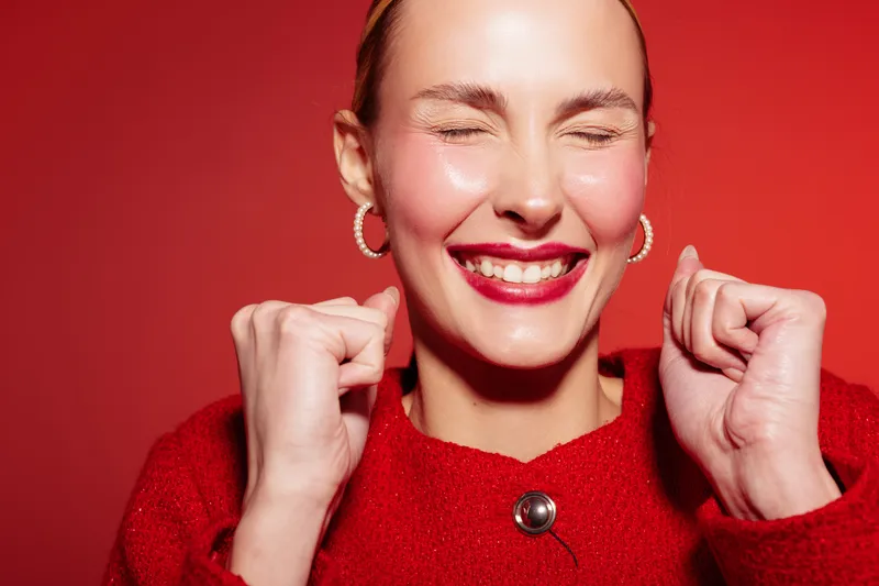 Happy woman in red sweater smiles with closed eyes and clenched fists on red background. Concept of joy, excitement, celebration, and positive emotions in studio portrait.
