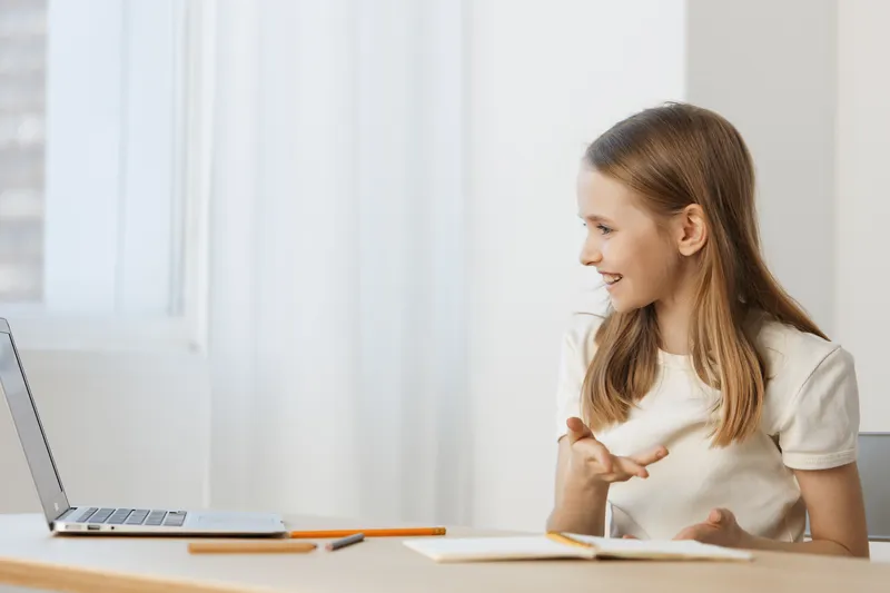 Young girl communicating online, expressing joy and engagement, sitting at a light desk with a laptop and notebook, bright and modern interior space