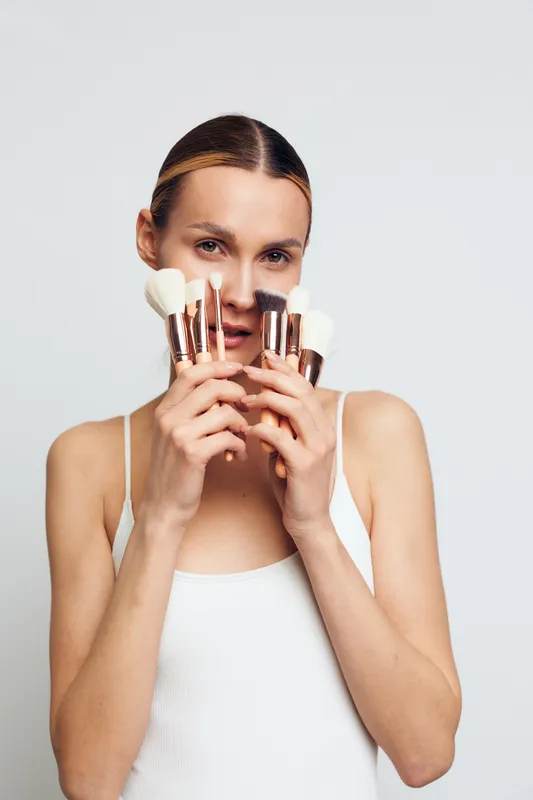 Young woman with smooth skin holding makeup brushes in front of her face on a white background. Concept of beauty, cosmetics, skincare, and personal care routine in studio light.