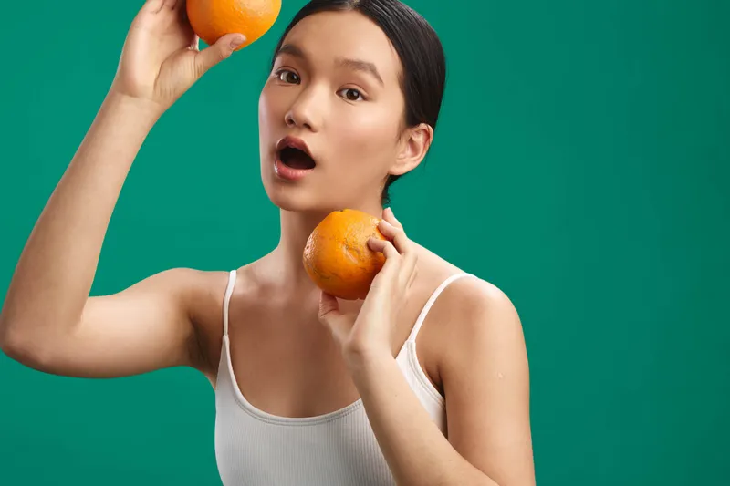 asian woman holding oranges against a vivid green background, casual sleeveless top, playful pose and surprised expression, creating a fresh, colorful lifestyle portrait.