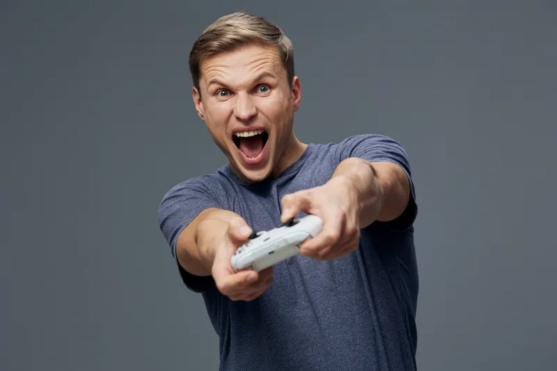 Excited young man with short hair in casual blue t-shirt passionately playing video game with game controller, expression of joy and concentration on his face, against plain gray background. People li
