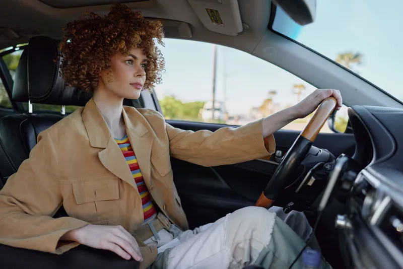 Young woman with curly hair drives a car, wearing a tan jacket and striped shirt, enjoying a calm moment with warm retro film color tones inside the vehicle