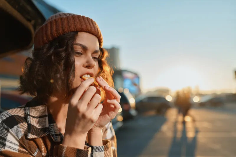 Woman on street eating fries at golden hour, candid lifestyle portrait with beanie and plaid, authenticity and emotional storytelling in an urban snack moment with warm golden hour glow.
