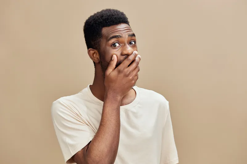Surprised Black man covering his mouth with his hand against a neutral beige background, expressing shock or disbelief, wearing a casual light colored t shirt