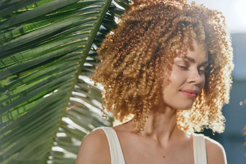 smiling woman with curly hair standing near tropical palm leaves wearing white top enjoying sunlight outdoors in warm weather