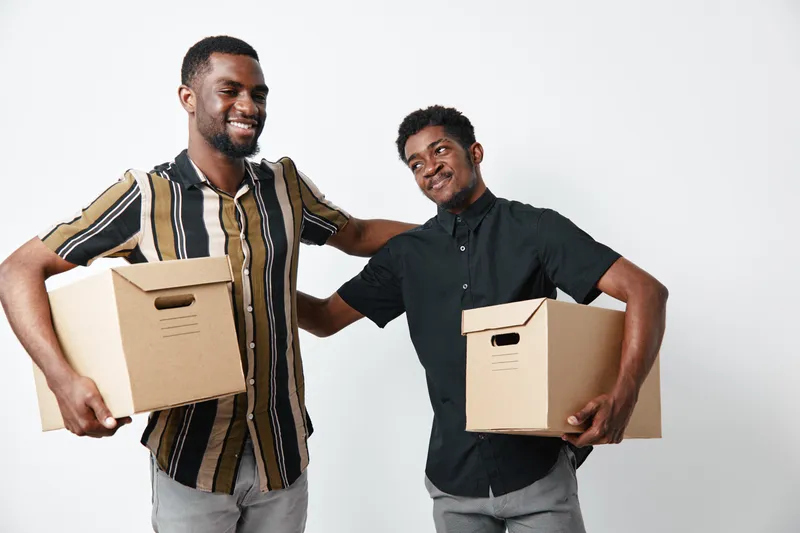 Two African American men smiling and holding cardboard boxes for moving or delivery. Young black males with dark skin in casual clothes on white background.